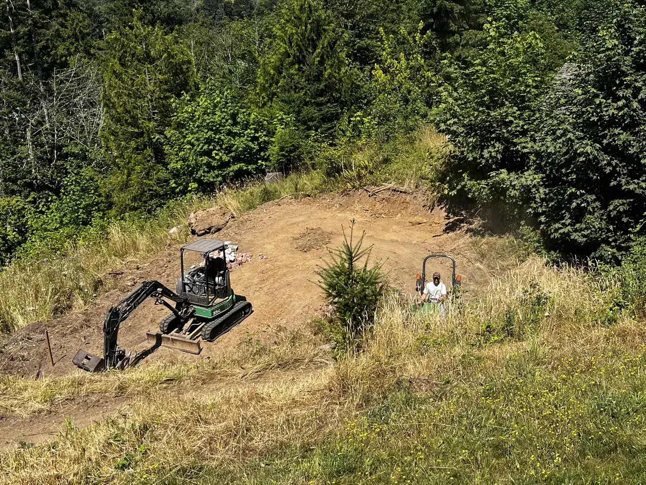A green mini excavator clearing a building pad on a grassy hillside